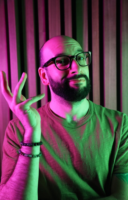 Connor’s headshot from mid-chest up. He is white, male, with short-buzzed head, neatly trimmed beard, and thick black-rimmed glasses. He looks directly at the camera, head slightly tilted, with an expression somewhere between asking a question and listening to an unexpected answer. His left arm lays across his lower chest (covered with a t-shirt) while his right hand is held up aloft in a “who knows?” pose. Right wrist has two beaded bracelets.